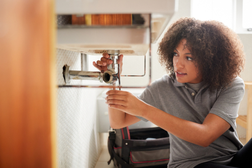 Young black female plumber sitting on the floor fixing a bathroom sink_ seen from doorway