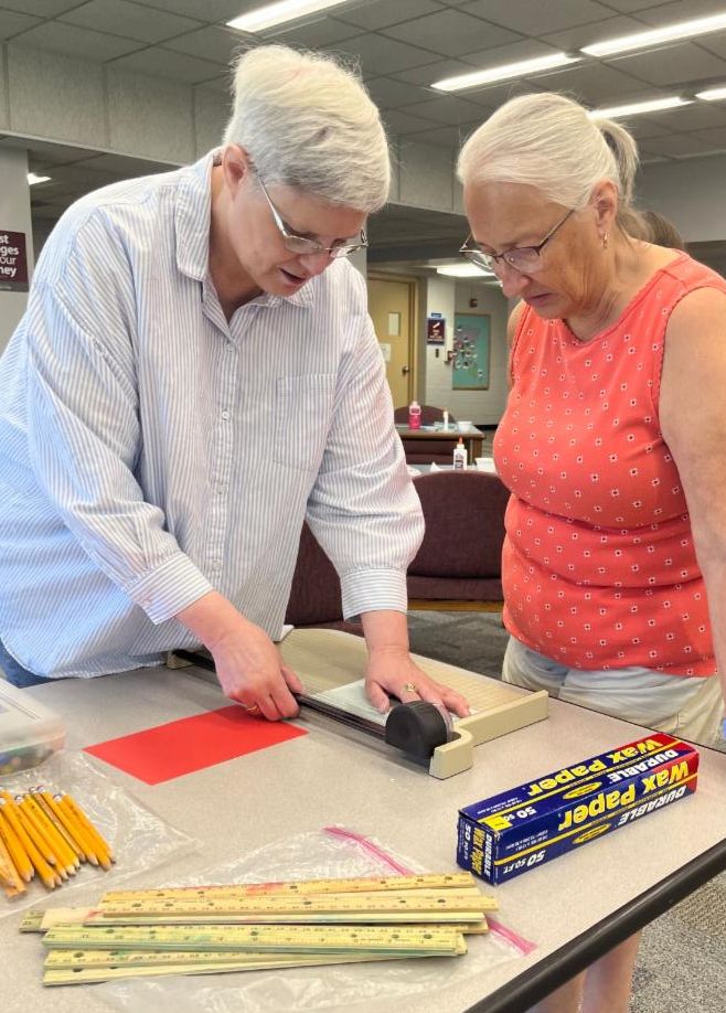 A women watches the instructor demonstrate with a page cutter at a table with rulers, pencils, and wax paper.