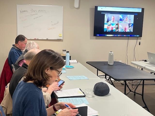 Several board members sit at tables while a television screen shows additional remote meeting goers.
