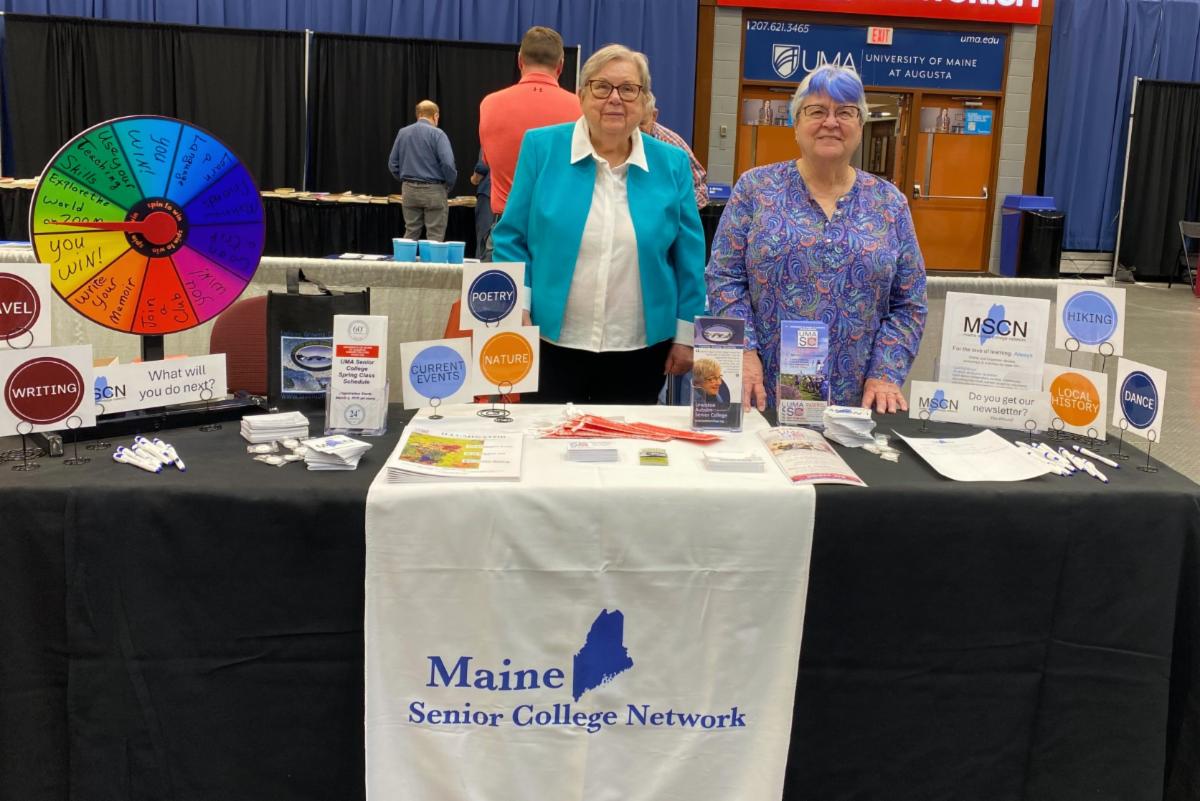 Two senior college volunteers stand behind the senior college event table with brochures and a prize wheel.