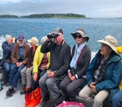 Members of Acadia Senior College sit along the side of a boat in the bay. One member uses binoculars.