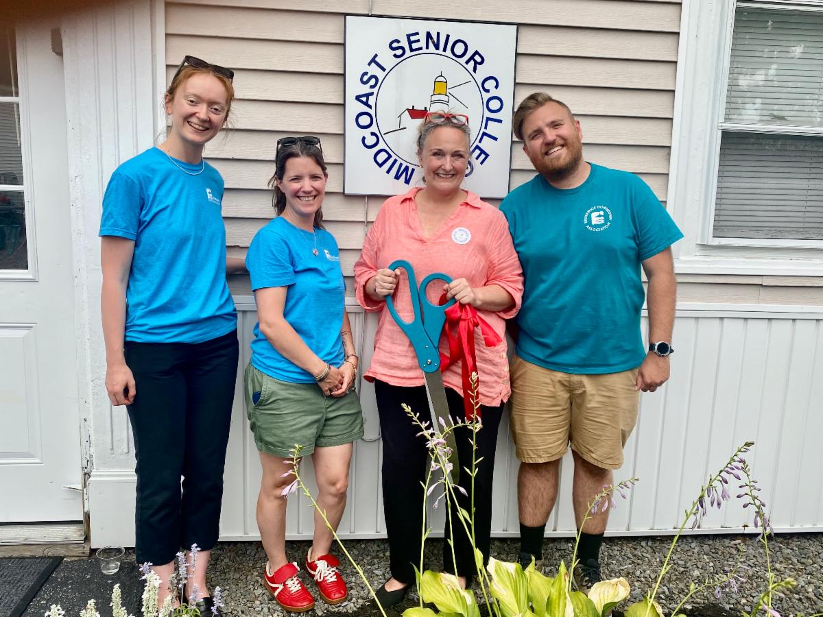 Midcoast Senior College director stands with three representatives from the Downtown Association holding a giant pair of scissors in front of the senior college building with its sign.
