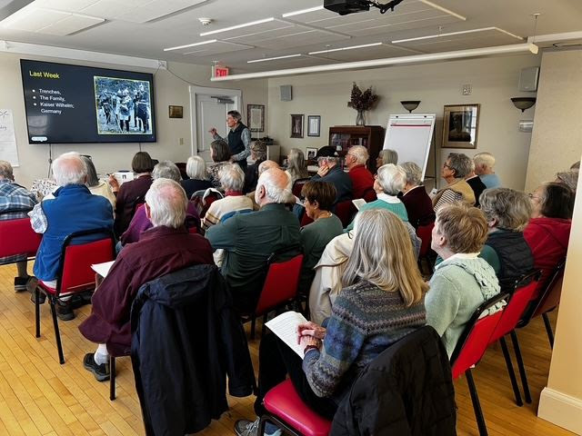 A large group of senior college members are seated and listening to a lecturer and looking at a slide.