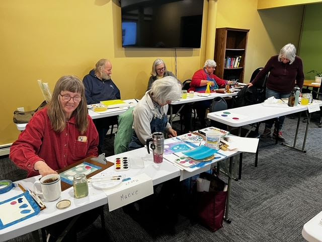 Six senior class members sit a long tables surrounded by watercolor paints, jars, and paper.