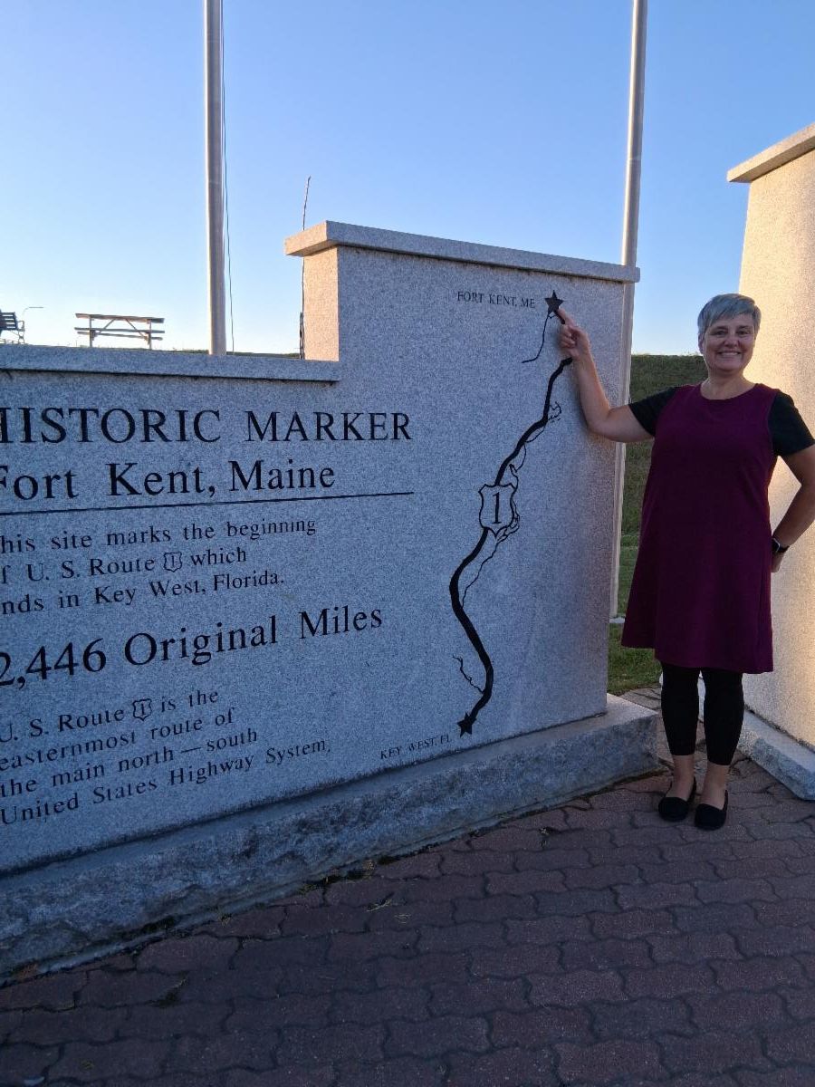 A woman stands beside a stone sign indicating the historic marker for Fort Kent, Maine, and points to a star at the top of a map of Route 1.