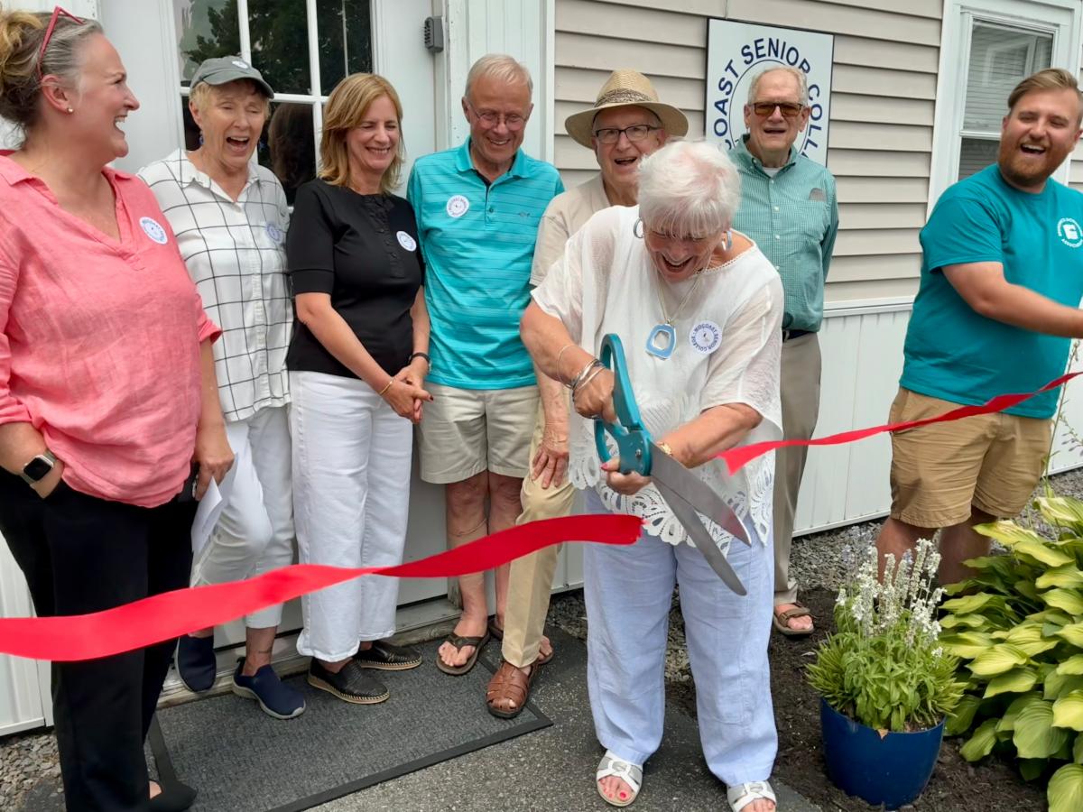 The Midcoast president cuts a ribbon with giant scissors while others gather around smiling and laughing in front of the new building.