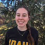 A young woman standing in front of a tree outside. She has long dark hair in a braid falling over her shoulder. She is smiling at the camera and wearing a black t-shirt with the word Idaho in bold yellow letters on it.