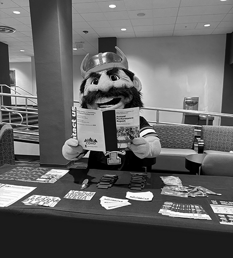 Joe Vandal sitting at a table with different bits of CDHD related swag in front of him. He is holding up a CDHD annual report.