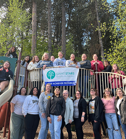 Women of different ages gathered together on a piece of playground equipment. They are all smiling and looking at the camera. There is a large banner at the center that reads IdahoSTARS Quality Achievers.