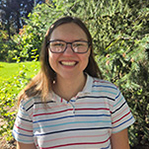 Emily is smiling and standing in front of a green leafy background. She is wearing a white top with multi color stripes and glasses. She has brown hair falling to her shoulders.