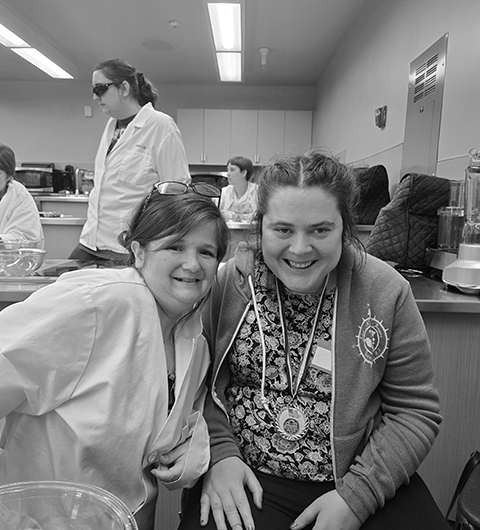 Two women sitting side by side. The woman on the left is leaning into the woman on the right. They are at a cooking station. Both are smiling happily and looking directly at the camera. 