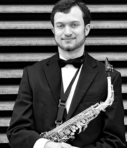 A young man smiles slightly at the camera. He is wearing a tuxedo and holding a saxophone.