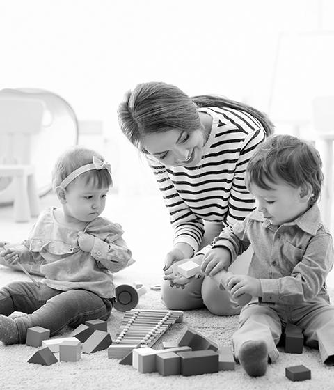 A female child care provider sitting with two toddlers on the floor, playing with baby toys.