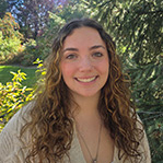 Natalie is smiling and standing in front of a green leafy background. She has long curly brown hair past her shoulders. She is wearing a tan cardigan over a white top.