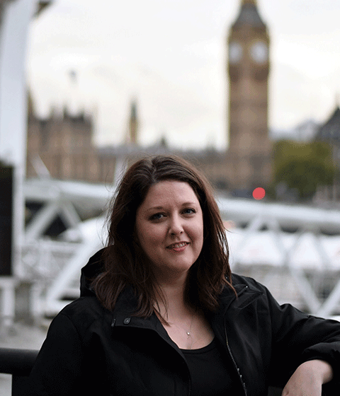A young woman leaning against a fence with Big Ben in the background. She is looking at the camera and smiling slightly.