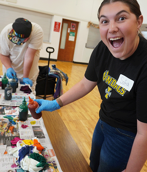 A young woman is smiling excitedly as she is holding a bottle of dye in her hand. She is working alongside self-advocates on a tie dye art project.