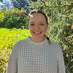Bella is smiling at the camera. She is standing in front of a green leafy background. She is wearing a white sweater and has shoulder length brown hair pulled back.