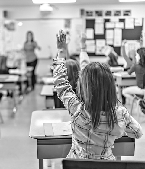 Looking to the front of an elementary classroom from behind. All the children are seated at desks, with many raising their hands. The teacher is at the front of the classroom.