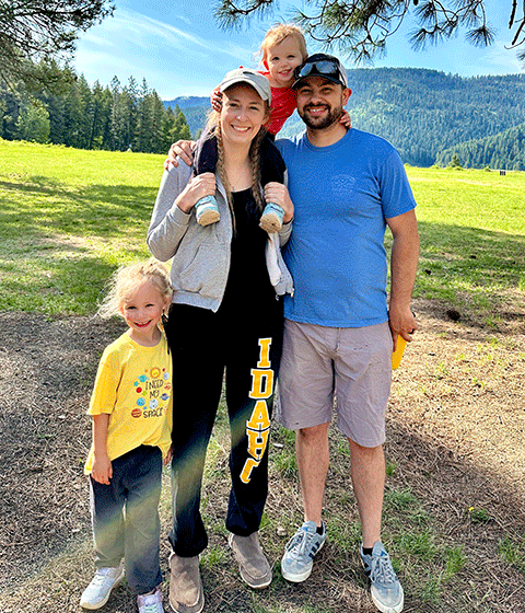 A young woman posing with her husband and two young children. They are all standing outside on a sunny day, smiling at the camera.