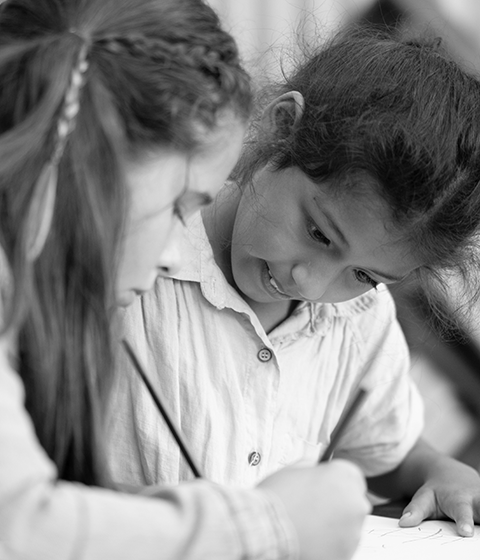 Two young girls are sitting together in a classroom. They are working on an assignment together. Both are looking at the paper intently while the girl on the left writes with a pencil. 