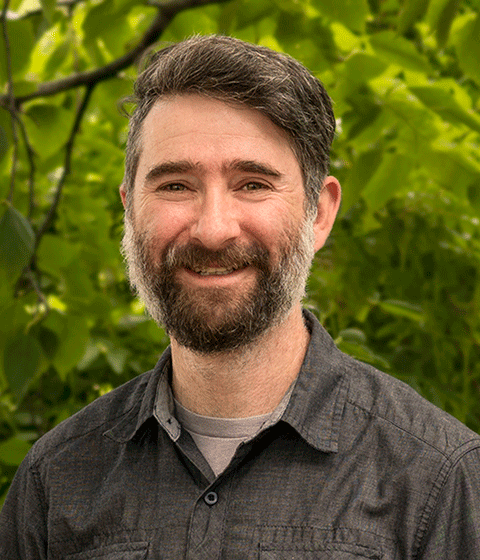 A man with dark hair and a salt and pepper beard is standing outside smiling at the camera. He is wearing a dark button-up shirt.