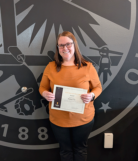 A woman wearing an orange top and dark pants, holding an award certificate. She is smiling at the camera.