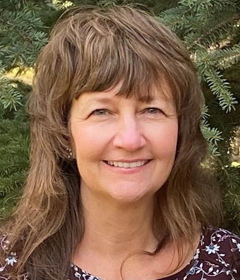 A woman standing outside in front of an evergreen. She is smiling slightly at the camera.