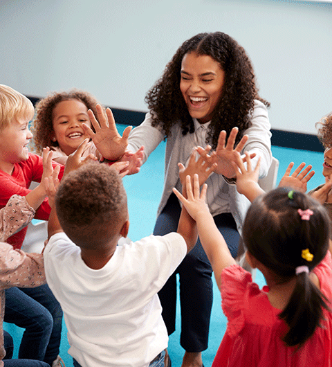 A teacher with her kindergarten students gathered around her. She is smiling and laughing as they all give her high fives. She is a person of color, with dark curly hair falling around her shoulders. Her students are boys and girls of different races.