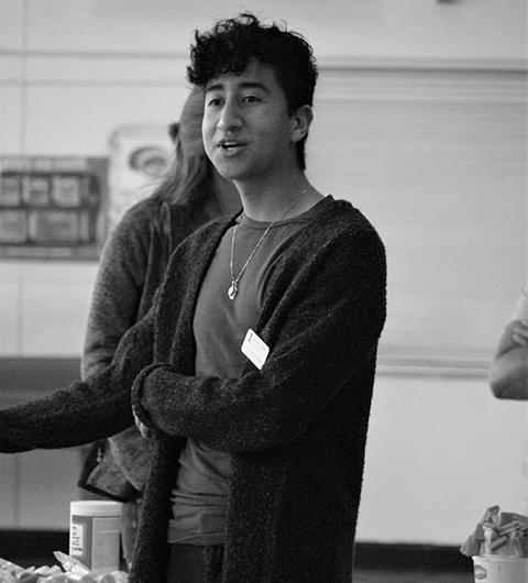 A young man standing with one of his arms across his body. He is addressing a group of people off camera. He has short dark hair and is wearing a dark cardigan over a dark t-shirt.