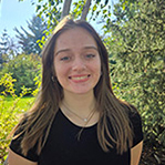 Hailey is smiling and standing in front of a leafy background. She is wearing a black top. Her long brown hair is falling around her shoulders.