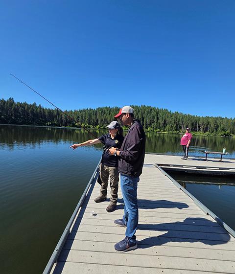 A woman pointing at the lakes surface as she helps a male self-advocate cast his fishing line into the lake.