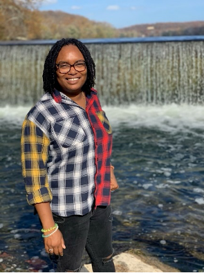 Kevina Brown is an African American female pictured smiling, with shoulder-length black hair and eyeglasses, dressed in a yellow, white, red, and black plaid shirt and black pants with a waterfall in the background