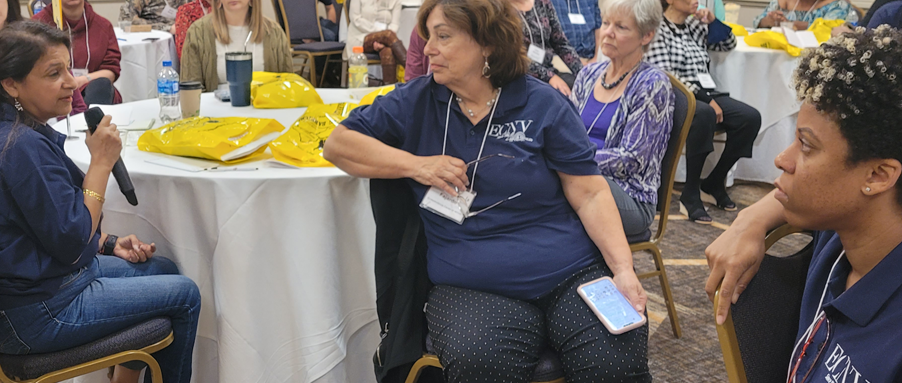 [Image: Director of Administration, Ruchika Lalwani takes the mic as Rosalia Fajardo (center in blue) and Dominique Dunford-Lack (far right in blue) listen]. 