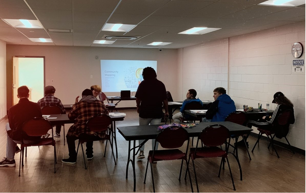 Seven mentees are sitting in a room at 4 different rectangle tables. Six mentees are facing an image projected on the wall at the front of the room while one mentee is turned around facing Kevina Brown, RAMP Program Coordinator, who is standing with her back to the camera in the middle of the mentees. 