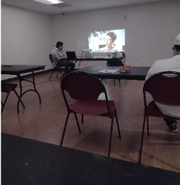 [Image: Students are seated in a classroom and watching an interactive video, Workplace Essential Skills Video: The Interview, that Is being projected onto the wall. One student is seated next to the projected video wearing a white sweater and assisting Kevina Brown (RAMP Program Coordinator)].