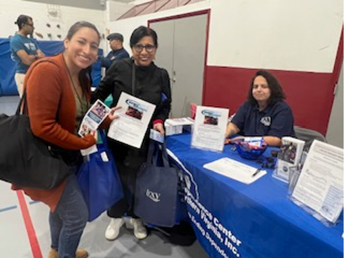 [Image: ​​Margarita Baquero (seated), giving away informative brochures and bags with COVID-19 home test to two Latina women attending the Fair]. 