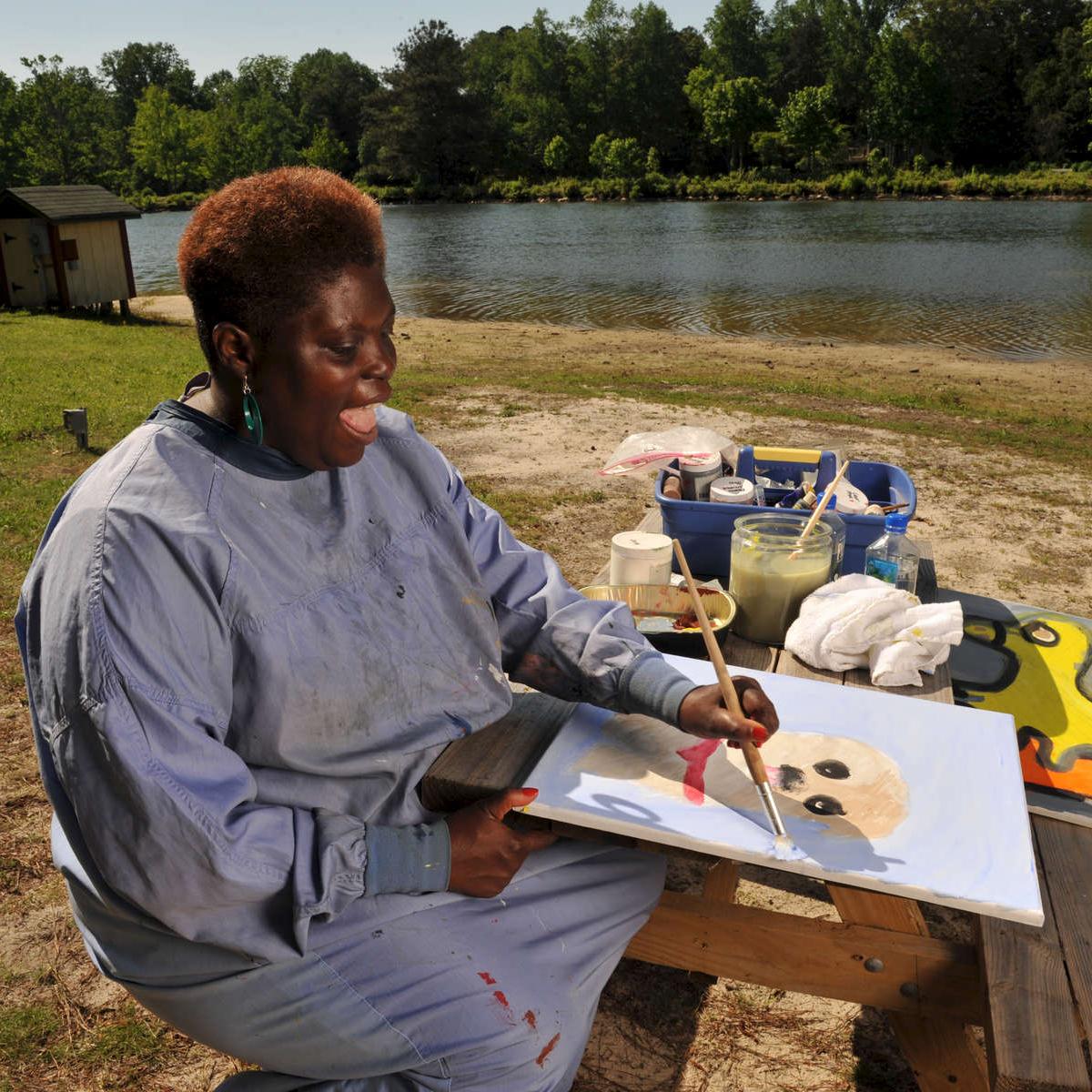 [Image: Lois Curtis, outside and near a lake, painting a portrait of a puppy with a red collar].