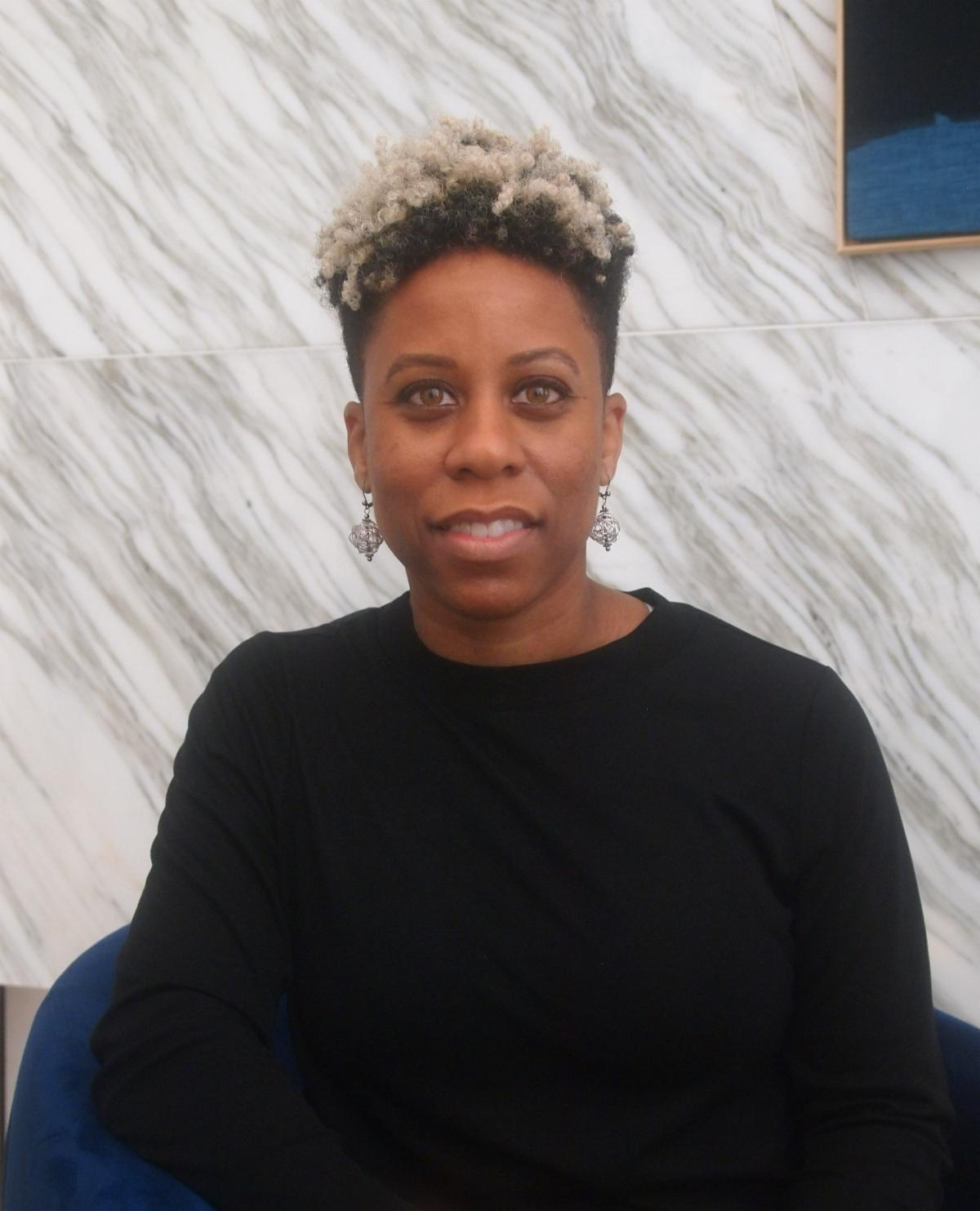 Dominique a black woman with short hair and a black longsleeve top and silver earrings sitting in a chair smiling for the camera