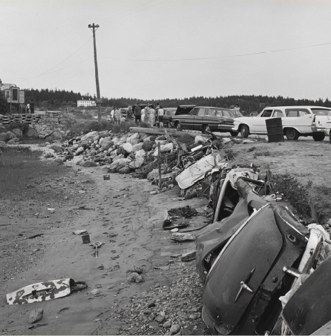 A black and white photograph shows old automobile parts strewn across a beach