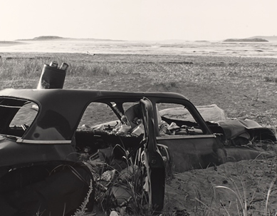 A black and white photograph of the remains of a discarded vehicle on a beach
