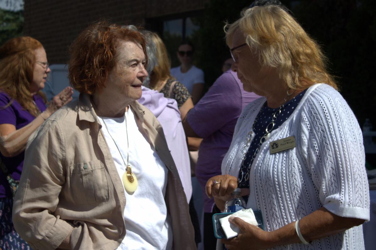 Two older women are standing and chatting with each other. The woman on the left has reddish short hair and is wearing a white shirt with a brown jacket. Her hands are in her pants pockets. The woman on the right has blond short hair and is wearing a white top. In the background, there are people mingling, but they are out of focus.