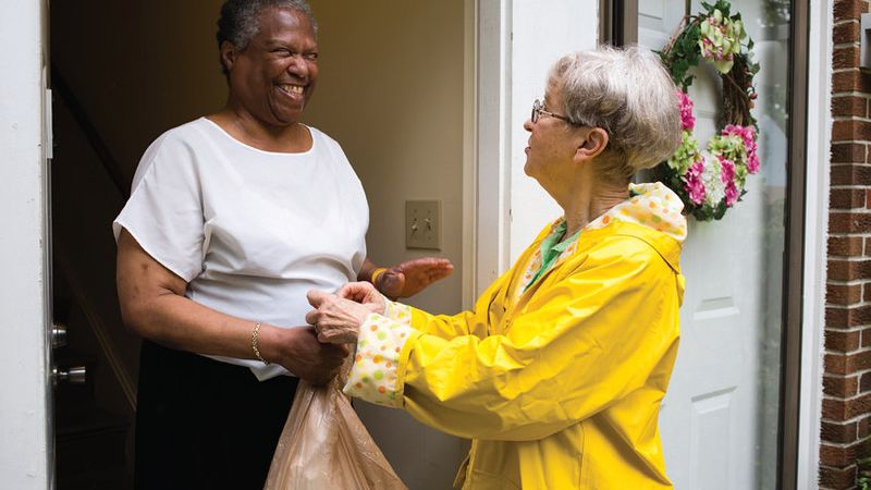 An older woman wearing a yellow raincoat smiles warmly as she hands a plastic grocery bag to another older woman standing in a doorway. The woman receiving the bag is smiling and appears to be expressing gratitude. A floral wreath hangs on the door, creating a welcoming atmosphere.