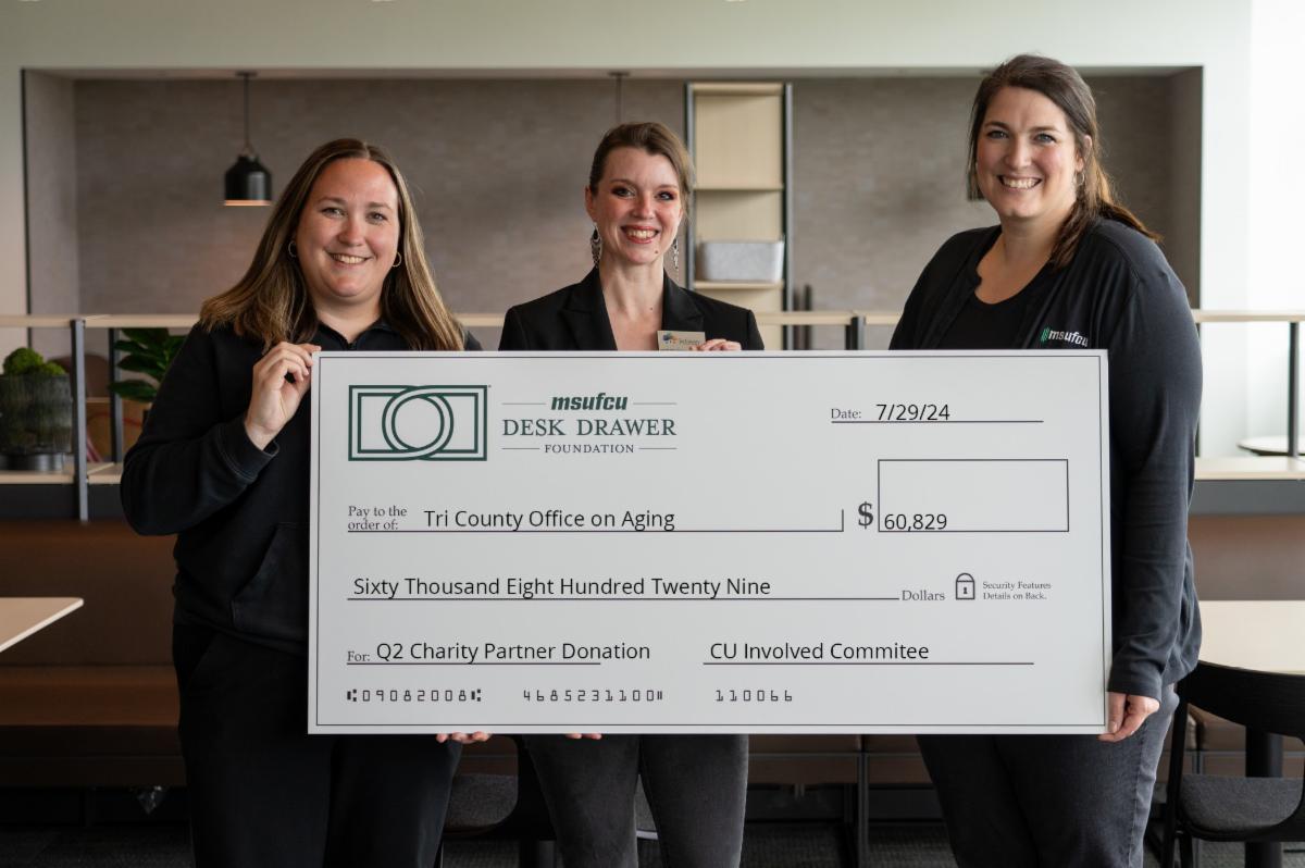 Three women are standing together, each holding a section of the giant check from MSUFCU's Desk Drawer Fund. They are all smiling broadly, radiating joy and accomplishment. The check displays the amount of $60,829, made out to Tri-County Office on Aging.