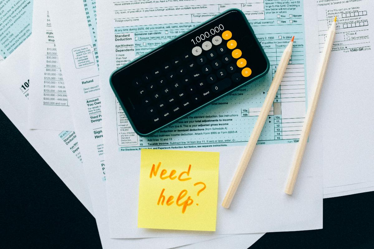 A stack of tax papers, pencils, a smartphone calculator, and a sticky note that says "Need help?"