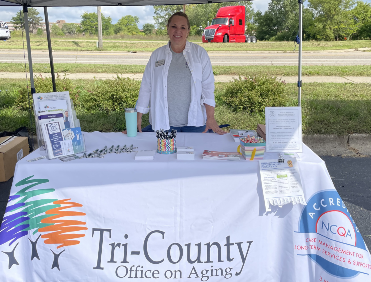  Karen Smith, a Tri-County Office on Aging (TCOA) employee, stands at an outreach table covered with a TCOA tablecloth. She engages with passersby, surrounded by colorful brochures showcasing the organization’s services for the aging community.