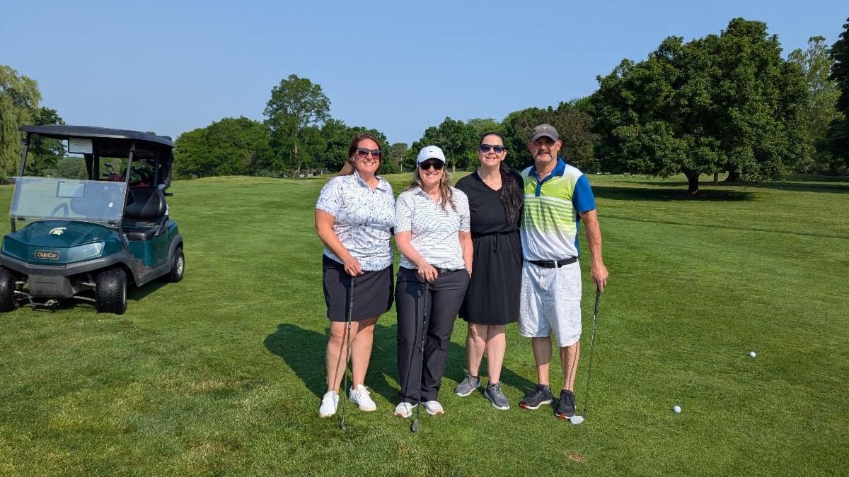 Four people stand together on a sunny golf course, smiling at the camera. Three women and one man are dressed in golf attire, holding golf clubs. A green golf cart is parked to the left, and golf balls are visible on the neatly mowed grass. Behind them, trees and a clear blue sky complete the background.