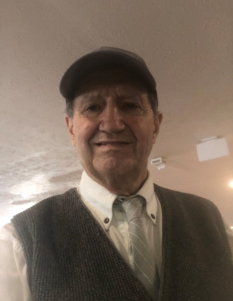 : A man stands wearing a brown hat and a brown vest. Underneath the vest, he has on a white collared shirt. 