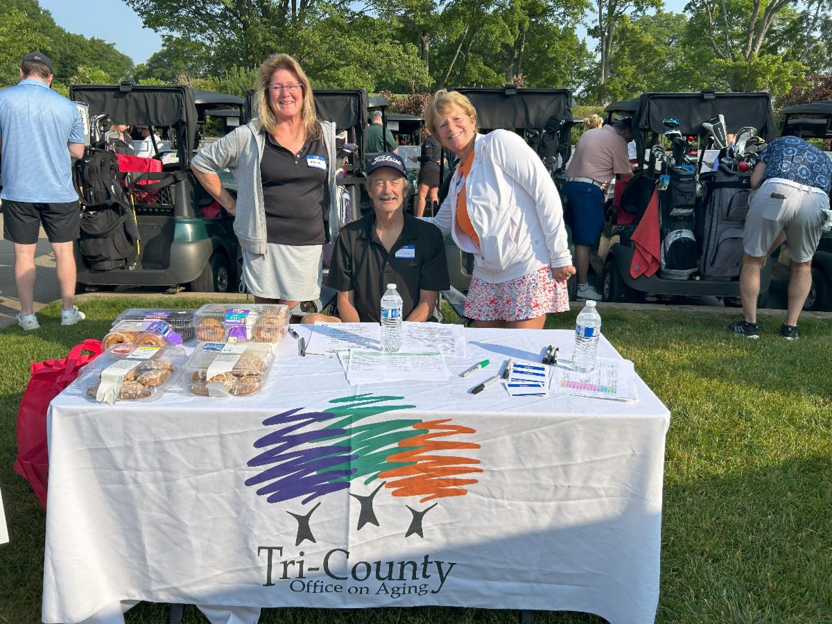 Three smiling adults stand and sit behind a registration table covered with a white Tri-County Office on Aging tablecloth. The table holds pastries, pens, papers, and water bottles. Behind them, golf carts and participants prepare for a golf event on a sunny day.