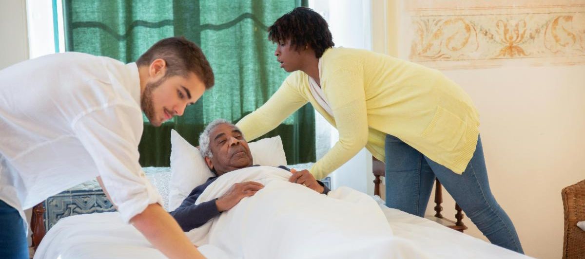 Two caregivers help an older man in a bed.