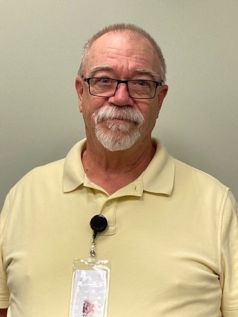 A man stands smiling with a friendly look on his face. He has a clean goatee and wears glasses. He’s dressed in a bright yellow polo shirt with a badge hanging from the collar. His smile and appearance show that he is approachable and kind.